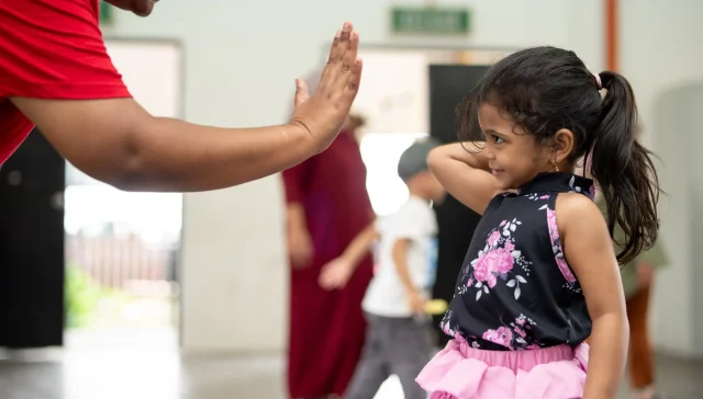 An NGO volunteer in Malaysia in red t-shirt high-fiving a little girl wearing pink skirt.