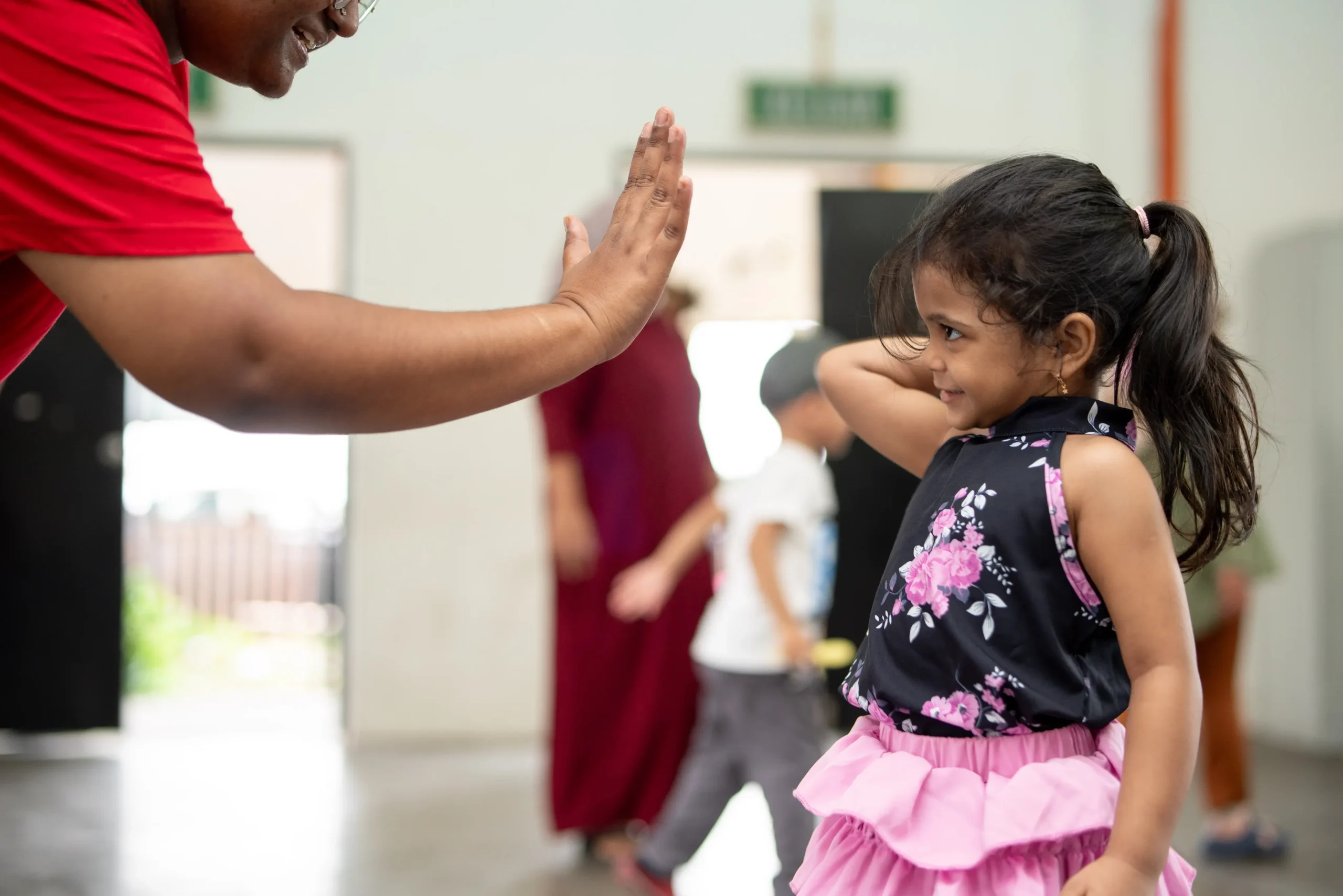 An NGO volunteer in Malaysia in red t-shirt high-fiving a little girl wearing pink skirt.