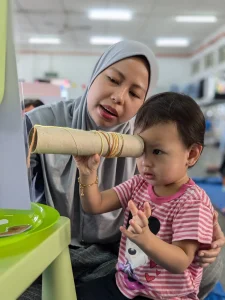 A boy looking through a cardboard spyglass next to his mother wearing gray hijab.