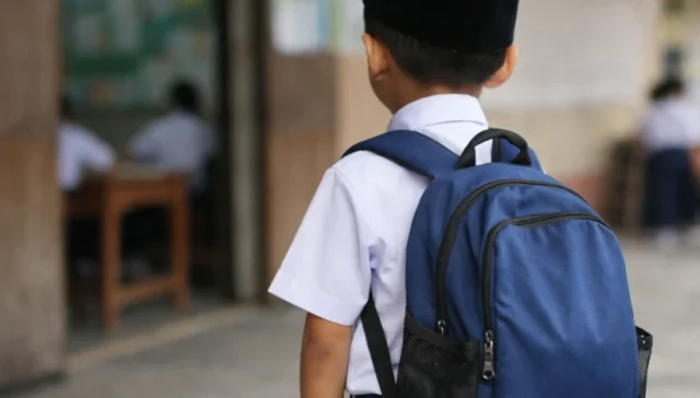 School readiness in Malaysia is more than wearing a uniform and entering a classroom, it is about a child feeling prepared, confident, and supported as they begin their journey into school. In this image, a young boy stands just outside a primary school classroom, seen from behind as he looks in, wearing a white school shirt, dark blue trousers, and a black songkok, with a large blue backpack resting on his shoulders. The lighting is soft and the background slightly blurred, while inside other students are already seated at their desks as he pauses at the doorway, capturing the transition into school where readiness is not just academic but also emotional and social.