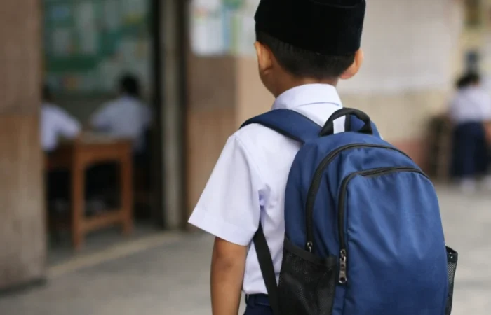 School readiness in Malaysia is more than wearing a uniform and entering a classroom, it is about a child feeling prepared, confident, and supported as they begin their journey into school. In this image, a young boy stands just outside a primary school classroom, seen from behind as he looks in, wearing a white school shirt, dark blue trousers, and a black songkok, with a large blue backpack resting on his shoulders. The lighting is soft and the background slightly blurred, while inside other students are already seated at their desks as he pauses at the doorway, capturing the transition into school where readiness is not just academic but also emotional and social.