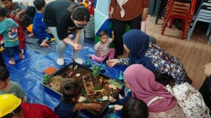 Children and caregivers gathered on a floor mat engaging in hands-on sensory play with a miniature setup of natural materials, toys, and tools. The scene shows a nurturing, interactive learning environment in Malaysia, reflecting community education and development initiatives supported through donation tax Malaysia contributions.